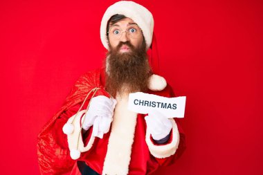 Uzun sakallı, Noel Baba kostümü giyen yakışıklı kızıl saçlı adam elinde noel mesajı ile kameraya bakıp öpüşmenin güzelliğine ve seksiliğine bakıyor. aşk ifadesi. 