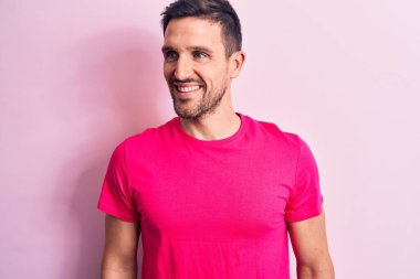 Genç yakışıklı bir adam, günlük tişört giyip pembe arka planda dikiliyor. Yan tarafa bakıyor, rahat bir görünüm takınıyor ve kendine güvenen bir gülümsemeyle gülümsüyor..