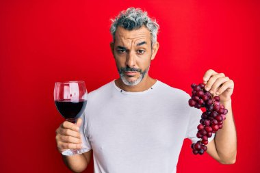 Orta yaşlı, gri saçlı, elinde taze üzüm dalı, kırmızı şarap kuşkucu ve gergin bir adam var. Sorunlar yüzünden surat asıyor. negatif kişi. 