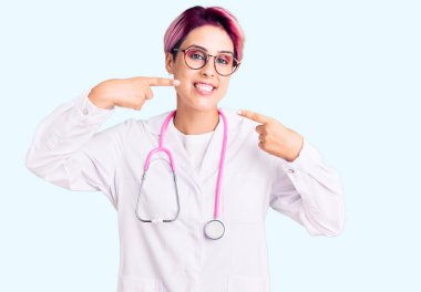 Pembe saçlı, doktor üniforması giyen neşeli bir şekilde gülümseyen ve parmaklarıyla ve ağzıyla işaret eden güzel bir kadın. diş sağlığı kavramı. 