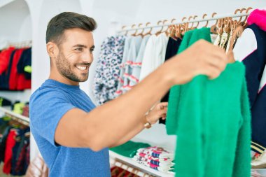 Genç, yakışıklı, gülümseyen, giyim mağazasında giysileri tutan.