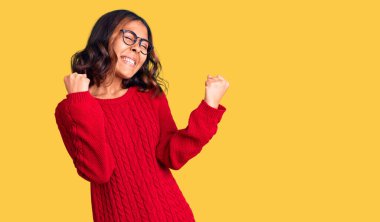 Kırmızı süveter ve gözlük takan genç ve güzel melez bir kadın çok mutlu ve heyecanlı kolları havada, gülümseyerek ve başarı için bağırarak kazanan bir jest yapıyor. kutlama konsepti. 