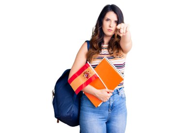 Güzel, genç, esmer bir değişim öğrencisi elinde İspanya bayrağı ile kamerayı işaret ediyor. Kendine güvenen bir jest, ciddi görünüyor. 