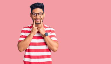 Yakışıklı Latin Amerikalı gündelik kıyafetler ve gözlük takan ağzı açık gülümseyen, parmakları işaret eden ve neşeli gülümsemeye zorlayan genç adam. 