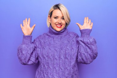 Güzel sarışın ve cüsseli bir kadın mor arka planda balıkçı yaka kazak giyiyor ve parmaklarını on numarayı gösterirken kendinden emin ve mutlu bir şekilde gülümsüyor..