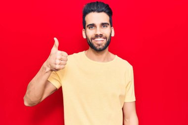 Sakallı, sıradan tişörtlü, mutlu ve pozitif gülümseyen, başparmağıyla mükemmel ve onay işareti yapan genç yakışıklı bir adam. 