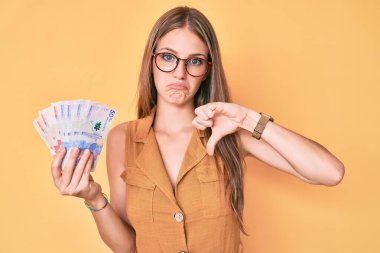 Elinde kızgın yüzlü Kolombiya pezosu tutan genç sarışın kız, olumsuz işaret baş parmakları aşağıda, reddedilme konsepti 