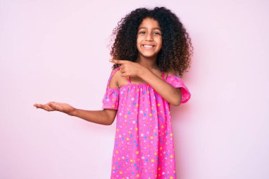 Kıvırcık saçlı Afro-Amerikalı çocuk, sıradan bir elbise giyip kameraya hayret ediyor ve gülümsüyor.. 