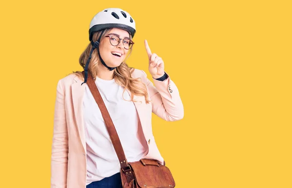 Young caucasian woman wearing bike helmet and leather bag pointing finger up with successful idea. exited and happy. number one.