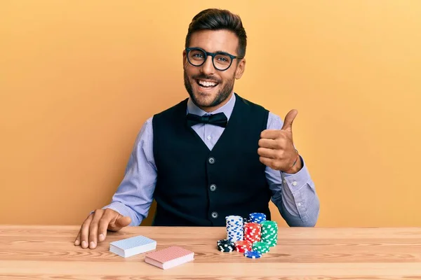 英俊而惊慌失措的男人坐在桌子上 拿着扑克牌和扑克牌开心而积极地微笑着 竖起大拇指做得很出色 签了字 — 图库照片