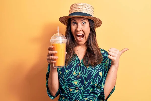 若いです美しいですブルネット女性身に着けています夏の帽子飲料ガラスのオレンジジュースポインティング親指へザ側笑顔幸せとともにオープン口 — ストック写真