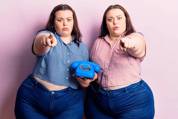 Young plus size twins holding vintage telephone pointing with finger to the camera and to you, confident gesture looking serious