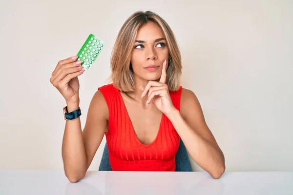 Beautiful caucasian woman holding birth control pills serious face thinking about question with hand on chin, thoughtful about confusing idea