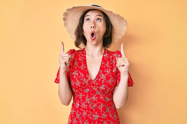 Yaz şapkası takan genç ve güzel kız şaşırmış ve şaşırmış bir şekilde yukarı bakıyor ve parmaklarıyla işaret ediyor ve kollarını kaldırıyor.. 