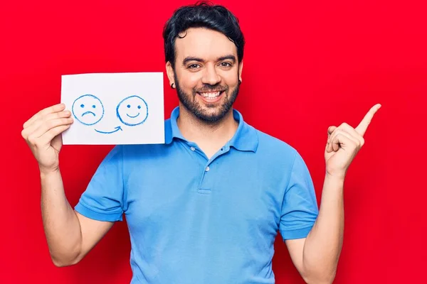 Young hispanic man holding sad to happy emotion paper smiling happy pointing with hand and finger to the side