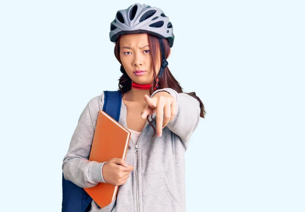 Young beautiful chinese girl wearing student backpack and bike helmet holding book pointing with finger to the camera and to you, confident gesture looking serious