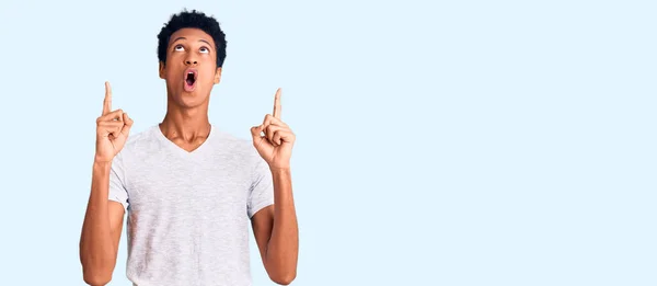 若いアフリカ系アメリカ人の男性は カジュアルな白いTシャツを着て驚きと驚きを見上げ 指と腕を上げて — ストック写真