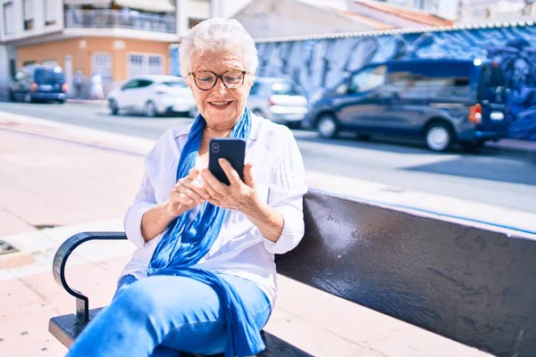 白髪の年配の女性が笑顔で屋外で幸せなスマートフォンを使用してベンチに座って — ストック写真