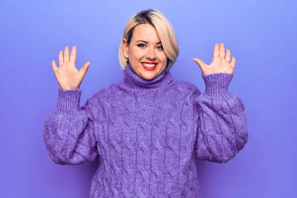Güzel sarışın ve cüsseli bir kadın mor arka planda balıkçı yaka kazak giyiyor ve parmaklarını on numarayı gösterirken kendinden emin ve mutlu bir şekilde gülümsüyor..