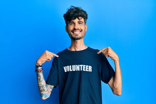 顔に笑顔で自信を持ってボランティアTシャツを着て若いヒスパニック系の男 誇りと幸せ指で自分自身を指して — ストック写真