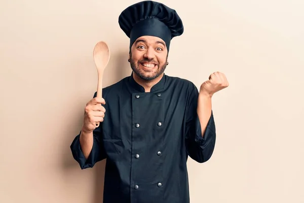 Young hispanic man wearing cooker uniform holding spoon screaming proud, celebrating victory and success very excited with raised arm