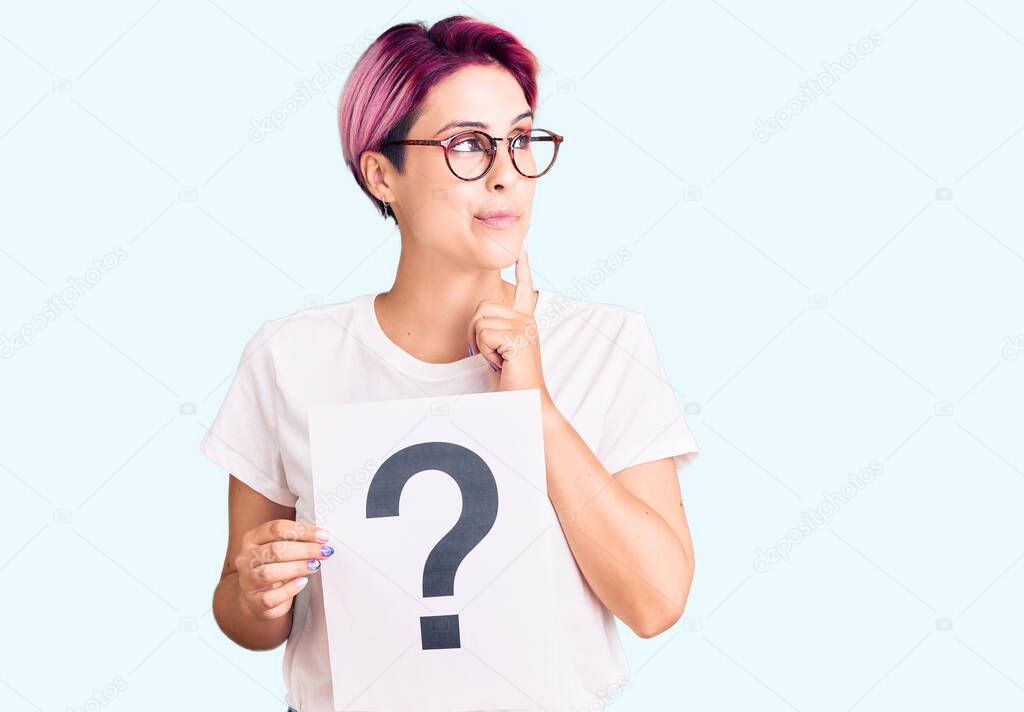 Young beautiful woman with pink hair holding question mark serious face thinking about question with hand on chin, thoughtful about confusing idea 