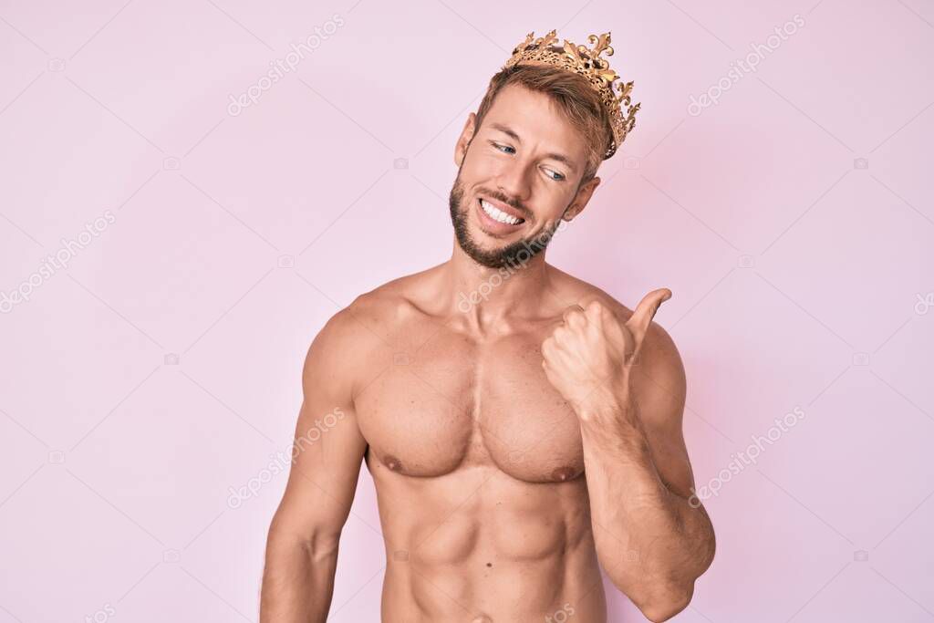 Young caucasian man shirtless wearing king crown smiling with happy face looking and pointing to the side with thumb up. 