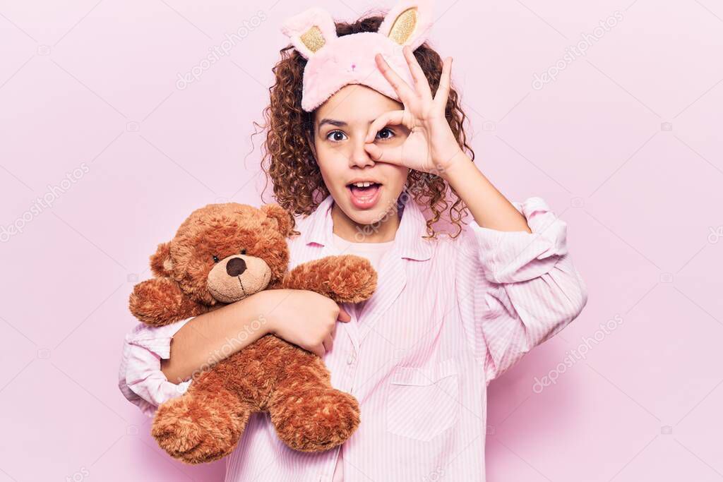 Beautiful kid girl with curly hair wearing sleep mask and pajamas holding teddy bear smiling happy doing ok sign with hand on eye looking through fingers 