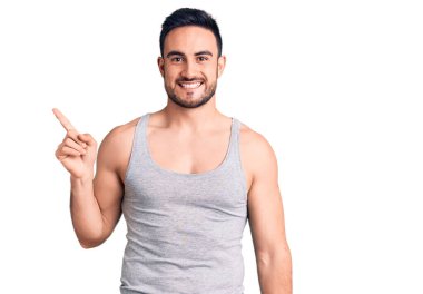 Mayo giyen, kolsuz tişörtlü, yüzünde kocaman bir gülümseme olan genç ve yakışıklı bir adam kameraya el ile işaret ediyor.. 