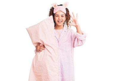 Kıvırcık saçlı, uyku maskesi takan ve yastık tutan güzel bir kız çocuğu. 