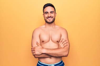 Sarı arka plan üzerinde kolsuz tişört giyen sakallı yakışıklı genç adam kameraya bakarken kollarını kavuşturup gülümsüyor. Pozitif kişi.