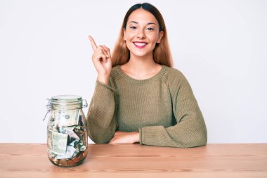 Masada içi dolar dolu bir kavanozla oturan genç esmer kadın mutlu bir şekilde elini ve parmağını kenarı işaret ederek gülümsüyor. 