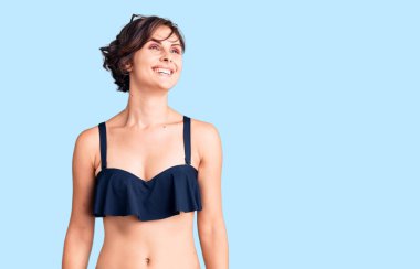 Kısa saçlı, bikini giyen, yüzünde gülümseme ve doğal ifadeyle yan yana bakan güzel bir kadın. Kendinden emin gülen. 