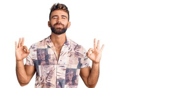 Latin kökenli genç adam yaz kıyafetleri giyip rahatlıyor ve gözleri kapalı gülümsüyor parmaklarıyla meditasyon hareketi yapıyor. yoga konsepti. 