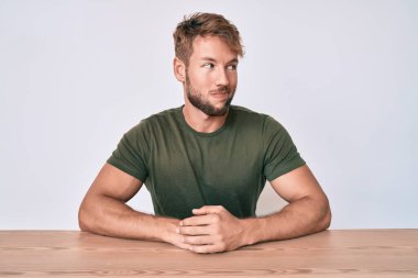 Gündelik giysiler giymiş genç beyaz bir adam masaya oturmuş gülümsüyor, yan tarafa bakıyor ve düşünüyor.. 