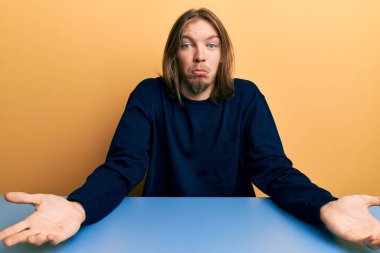 Masada oturan, uzun saçlı, sıradan kıyafetler giyen, elleri ve kolları havada, kafası karışık, yakışıklı, beyaz bir adam. şüphe kavramı. 