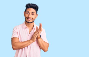 Yakışıklı Latin Amerikalı genç adam, sıradan bir yaz tişörtü giyiyor, alkışlıyor, alkışlıyor, mutlu, mutlu ve gururlu elleri birleştiriyor. 