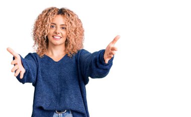 Kış süveteri giyen kıvırcık saçlı genç sarışın kadın kameraya sarılıp gülümsüyor. Mutluluğu kucaklayan neşeli bir ifade.. 