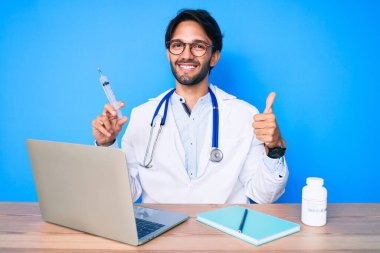 Klinikteki yakışıklı İspanyol doktor elinde şırıngayla mutlu ve pozitif gülümsüyor, başparmağıyla mükemmel ve onay işareti yapıyor. 