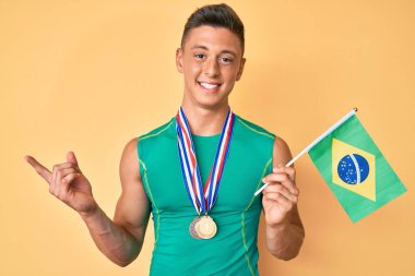 Spor kıyafetleri giyen ve Brezilya bayrağı tutan genç İspanyol çocuk mutlu bir gülümsemeyle elini ve parmağını yana doğru uzattı. 
