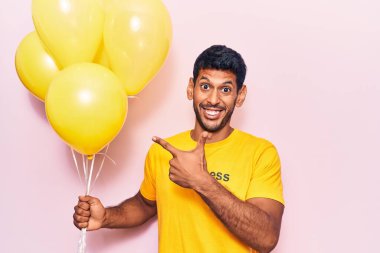 Üzerinde mutluluk mesajı olan tişört giyen genç latin adam elinde balonlar tutarken mutlu mesut elini ve parmağını gösteriyor. 