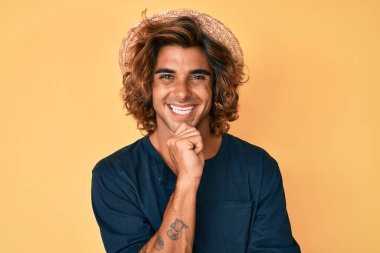 Yaz şapkası takan İspanyol genç adam kameraya bakıyor kollarını kavuşturup gülümseyerek ve çenesini kaldırarak. Olumlu düşünmek. 