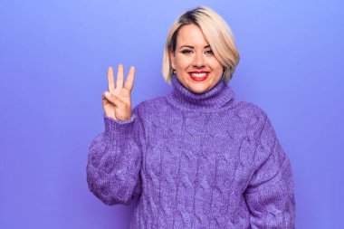 Güzel sarışın ve cüsseli bir kadın mor arka planda balıkçı yaka kazak giyiyor ve üç numaralı parmaklarıyla gülümseyerek mutlu ve kendinden emin görünüyor..