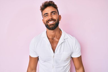 Gündelik kıyafetler giyen genç İspanyol adam pozitif ve mutlu görünüyor. Kendine güvenen bir gülümsemeyle ayakta duruyor ve dişlerini gösteriyor. 