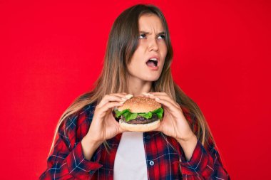 Güzel, beyaz bir kadın lezzetli klasik bir burger yiyor kızgın ve öfkeli çığlıklar atıyor öfkeyle bağırıyor. öfke ve agresif konsept. 