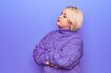 Güzel sarışın ve cüsseli bir kadın mor arka planda balıkçı yaka kazak giyiyor kolları çapraz ve kendinden emin bir şekilde yana bakıyor.