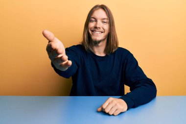 Uzun saçlı yakışıklı beyaz adam, sıradan giysiler içinde masada oturmuş dostça gülümseyerek selamlaşıp selamlıyor. başarılı iş. 