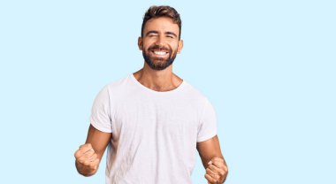 Gündelik beyaz tişört giyen genç İspanyol adam çok mutlu ve heyecanlı kolları havada, gülümseyen ve başarı için çığlık atan kazanan bir jest yapıyor. kutlama konsepti. 