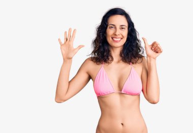 Genç ve güzel İspanyol kadın bikini giyiyor. Altıncı parmaklarını gösterip işaret ederken kendinden emin ve mutlu gülümsüyor.. 