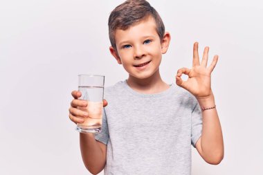 Sevimli sarışın çocuk bir bardak su içiyor. Parmaklarıyla işaret veriyor, gülümsüyor. Mükemmel bir sembol. 
